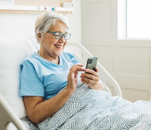Senior woman smiling while using smartphone in hospital bed wearing blue shirt.