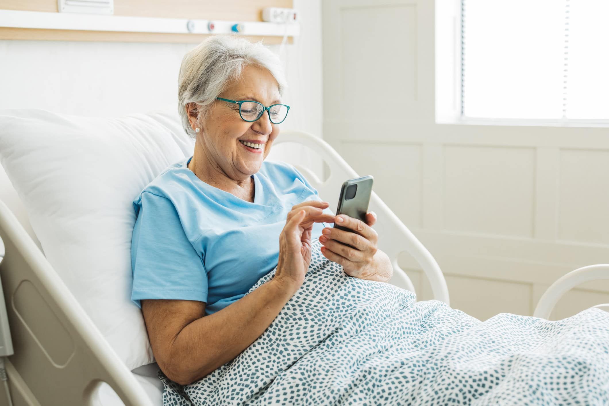 Senior woman smiling while using smartphone in hospital bed wearing blue shirt.