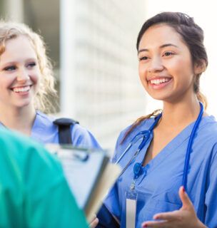 Young adult Hispanic woman is talking with young adult Caucasian blonde woman and young adult African American woman outside on college campus. Women are nursing or medical students. They are wearing hospital scrubs and stethoscopes.