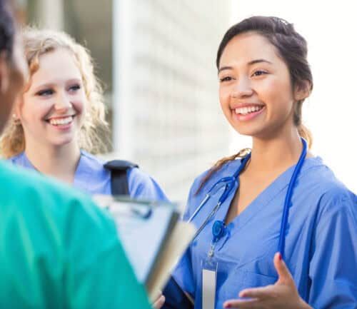Young adult Hispanic woman is talking with young adult Caucasian blonde woman and young adult African American woman outside on college campus. Women are nursing or medical students. They are wearing hospital scrubs and stethoscopes.