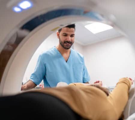 Man nurse looking at senior female patient lying on MRI scanner. Male medical technician operating CAT scan machine in hospital.