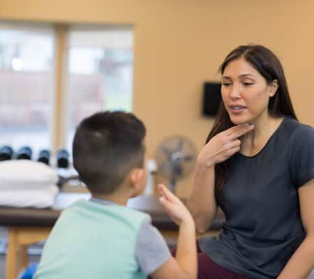 A speech therapist of Asian descent works with a handsome young elementary-age boy on his pronunciation. They are sitting on a treatment table with their forefingers pointing to their mouths and practicing the correct way of forming sounds and syllables. The shot is focused on her and over his right shoulder.