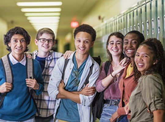 A multi-ethnic group of six high school students, 15 to 18 years old, hanging out together in the hallway by the lockers, between classes. There are three teenage boys and three teenage girls, smiling at the camera.