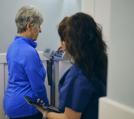 A nurse in a doctor’s office, wearing a senior female patient on a scale.