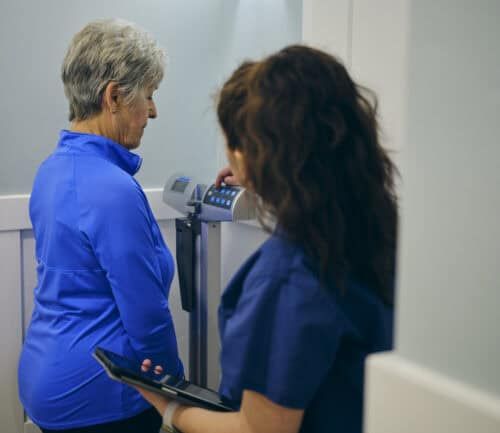 A nurse in a doctor’s office, wearing a senior female patient on a scale.