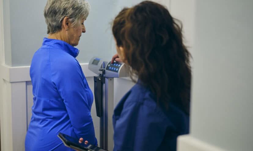 A nurse in a doctor’s office, wearing a senior female patient on a scale.