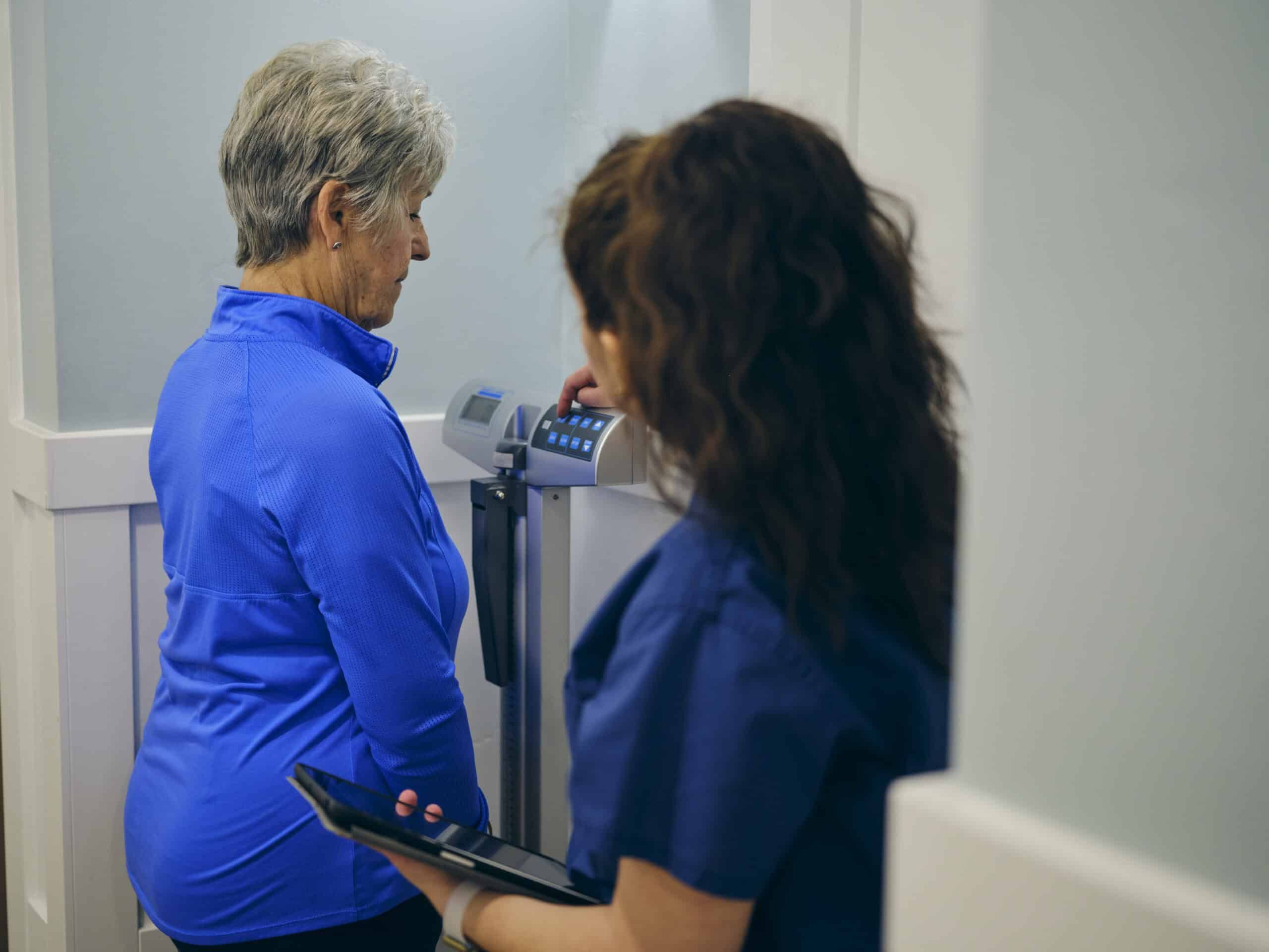 A nurse in a doctor’s office, wearing a senior female patient on a scale.
