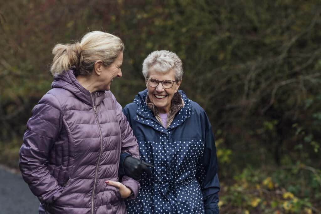 A mature woman walking with her mother on a path through a public park while getting exercise in Seghill, Northumberland. The senior woman has dementia and is arm in arm with her daughter for support and care while they look at each other, laugh and enjoy the fresh air.