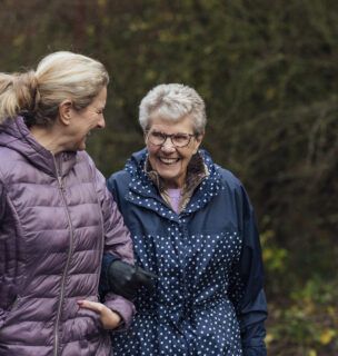A mature woman walking with her mother on a path through a public park while getting exercise in Seghill, Northumberland. The senior woman has dementia and is arm in arm with her daughter for support and care while they look at each other, laugh and enjoy the fresh air.