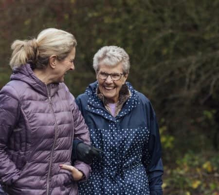 A mature woman walking with her mother on a path through a public park while getting exercise in Seghill, Northumberland. The senior woman has dementia and is arm in arm with her daughter for support and care while they look at each other, laugh and enjoy the fresh air.
