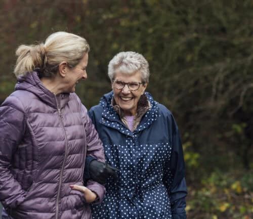 A mature woman walking with her mother on a path through a public park while getting exercise in Seghill, Northumberland. The senior woman has dementia and is arm in arm with her daughter for support and care while they look at each other, laugh and enjoy the fresh air.