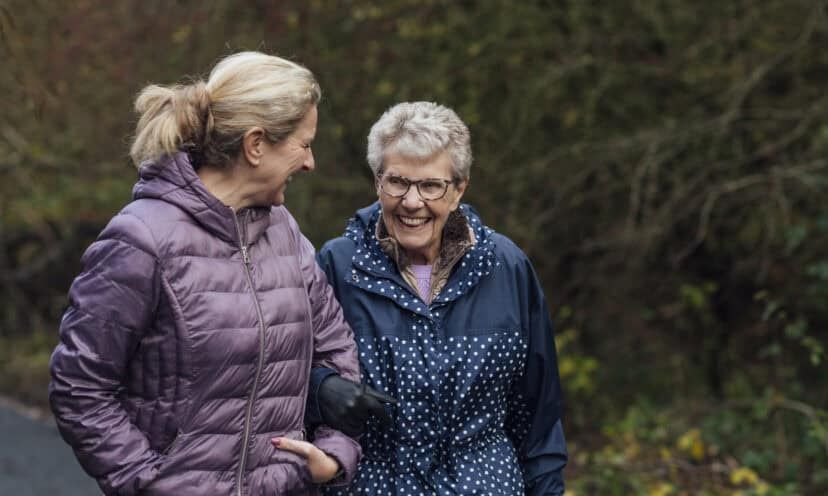 A mature woman walking with her mother on a path through a public park while getting exercise in Seghill, Northumberland. The senior woman has dementia and is arm in arm with her daughter for support and care while they look at each other, laugh and enjoy the fresh air.