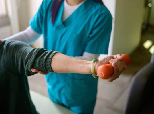 Female physiotherapist helping her senior patient raise dumbbells in a rehabilitation center.
