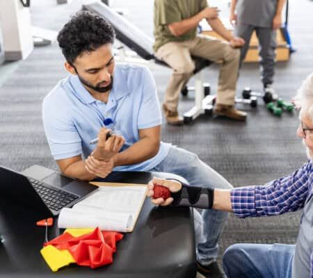 The young adult male physical therapist holds his own wrist as he discusses wrist injuries with the senior adult man.