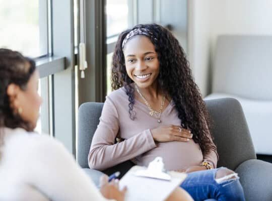 The expectant young adult woman smiles as she listens to advice from her unrecognizable female counselor.