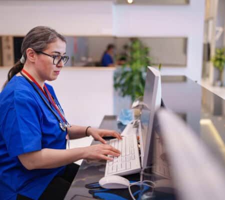 A healthcare worker is seen working at a reception desk in a medical facility, managing patient inquiries or administrative tasks. The scene highlights professionalism and the essential role of reception staff in healthcare settings.
