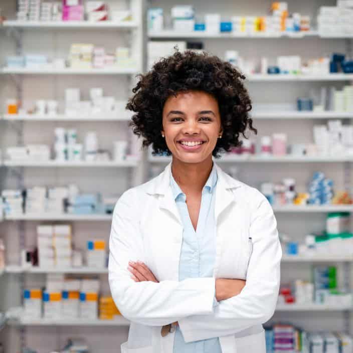 Confident female pharmacist wearing a lab coat, posing with arms crossed in a well-stocked pharmacy. Background features shelves filled with medicine, representing healthcare, professionalism, and a trustworthy environment.