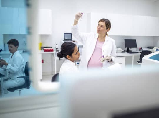 Wide-angle shot showing pregnant Caucasian scientist and young Indian female colleague analyzing test samples in bright, modern laboratory environment. Professional interaction during medical research.