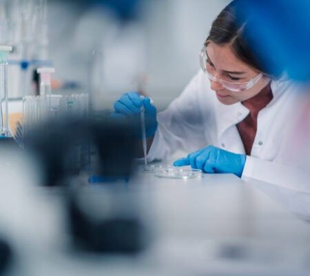 Young adult female scientist wearing safety glasses and blue gloves, carefully pipetting liquid into a petri dish during an experiment