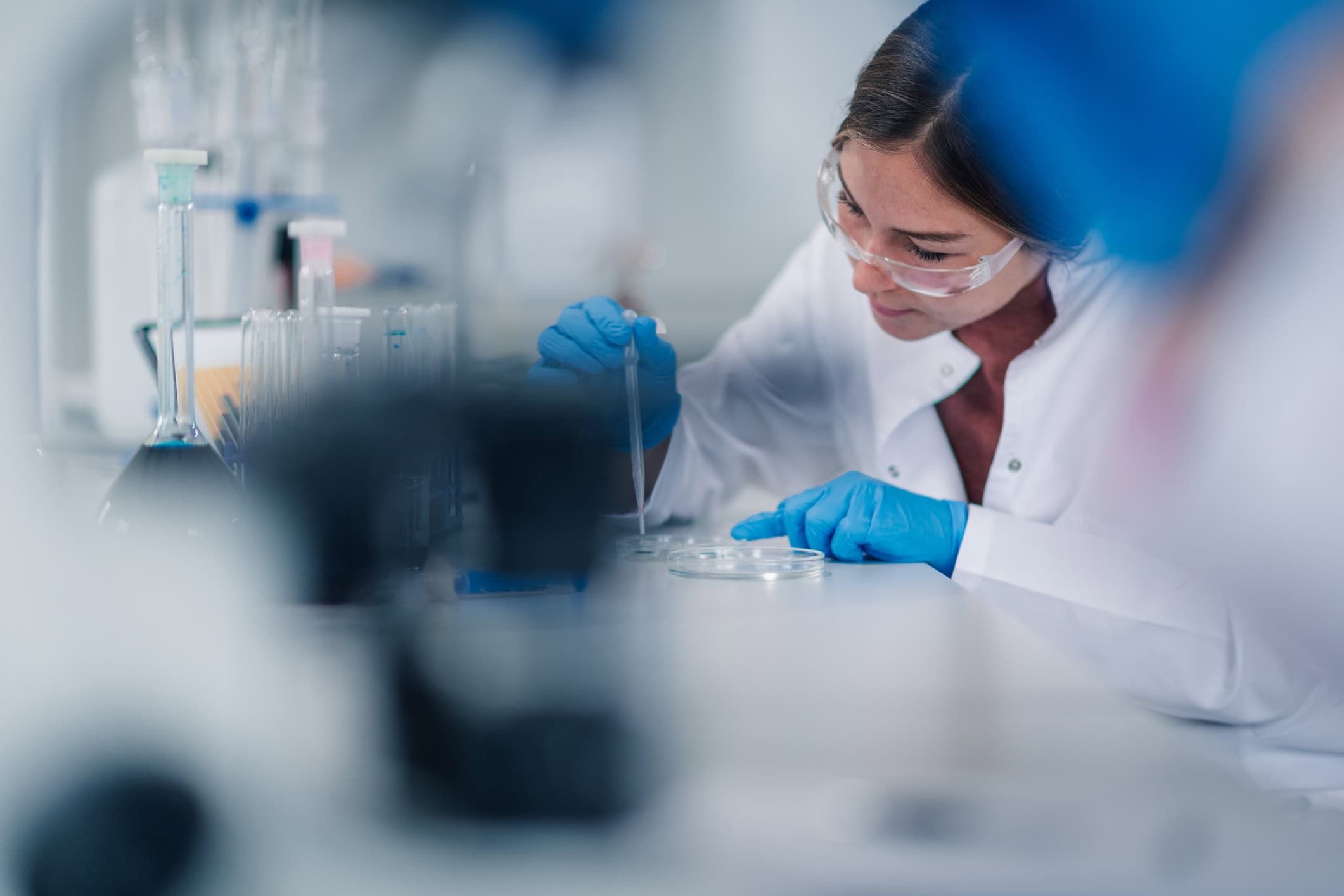 Young adult female scientist wearing safety glasses and blue gloves, carefully pipetting liquid into a petri dish during an experiment