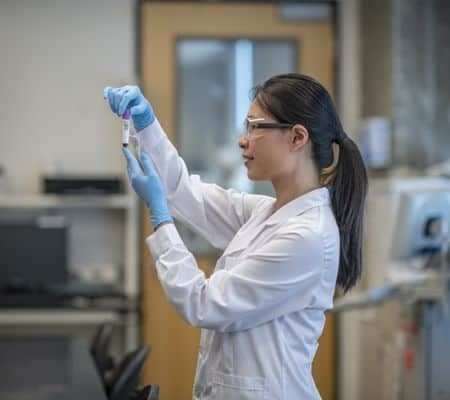 A female lab technician of Asian decent, holds up a vial of blood and looks at it closely before putting it into the centrifuge for processing.